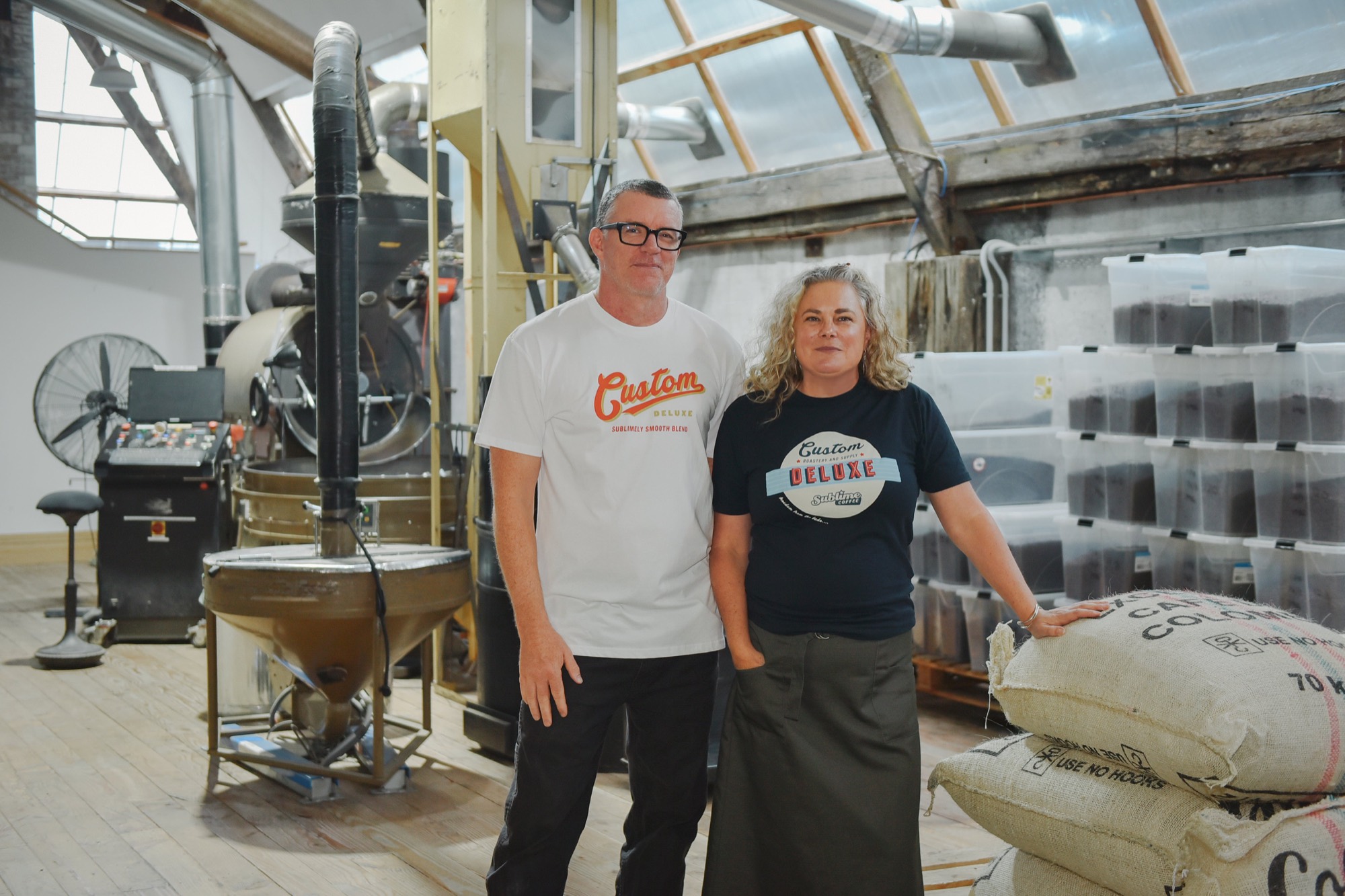 Dan and Emma Hennah standing in the Bridge Street roastery in front of the roaster and sacks of green coffee